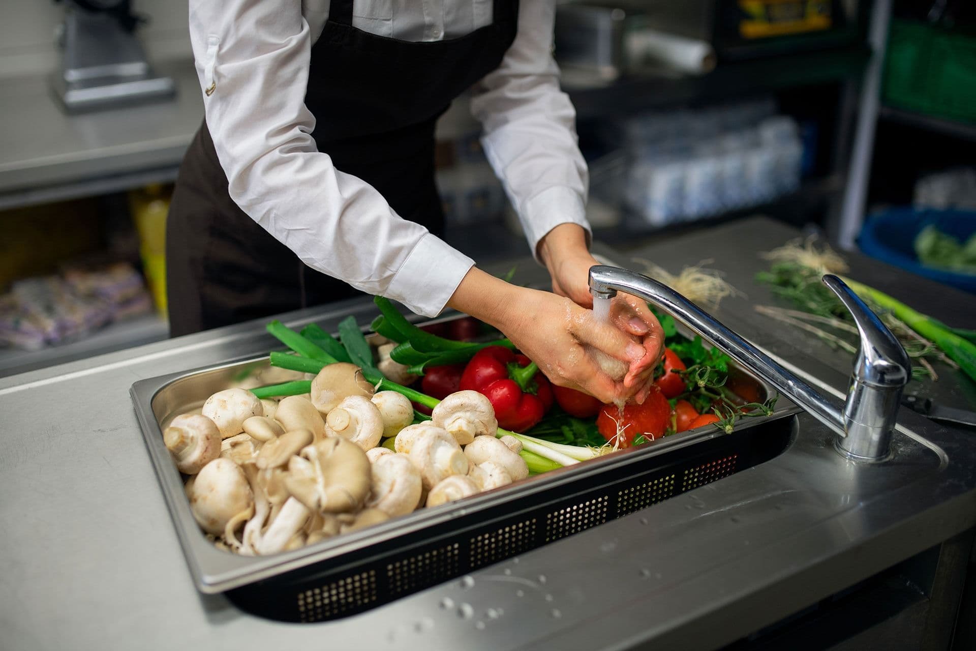 A person in an apron washes a variety of fresh vegetables, including mushrooms, green onions, and peppers, in a commercial kitchen sink.