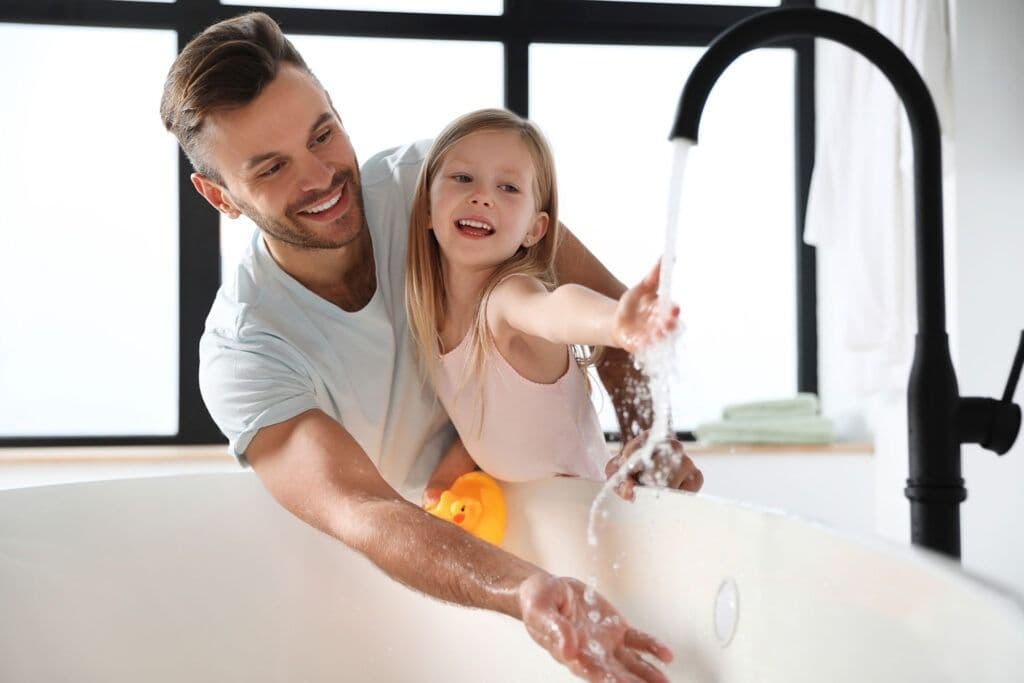 Father and daughter share a joyful moment at the bathtub, smiling as they play with running water. A rubber duck is visible, enhancing the playful scene.