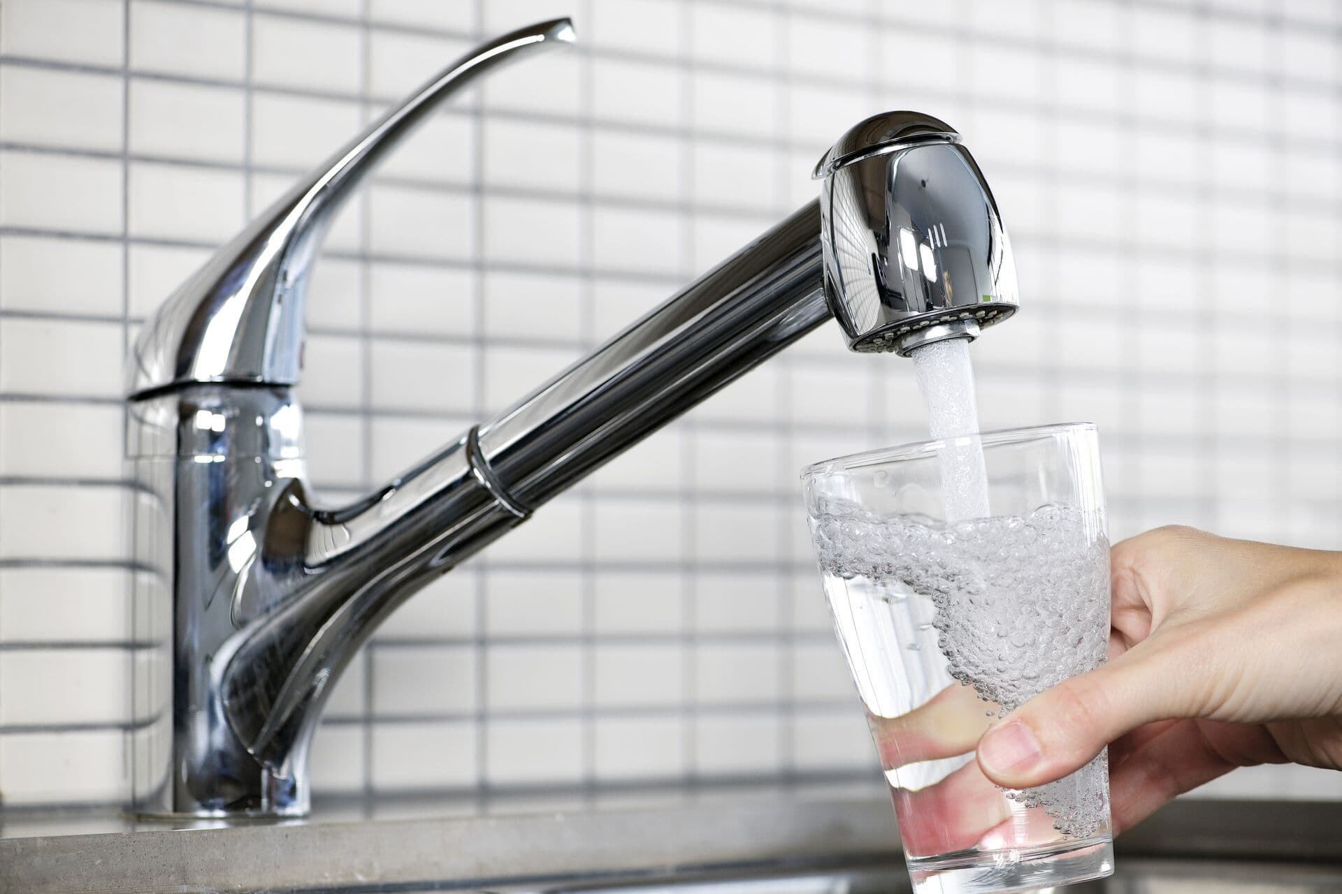 A hand holds a clear glass under a silver kitchen faucet, filling it with running water. The background features white tiled walls, evoking a clean setting.