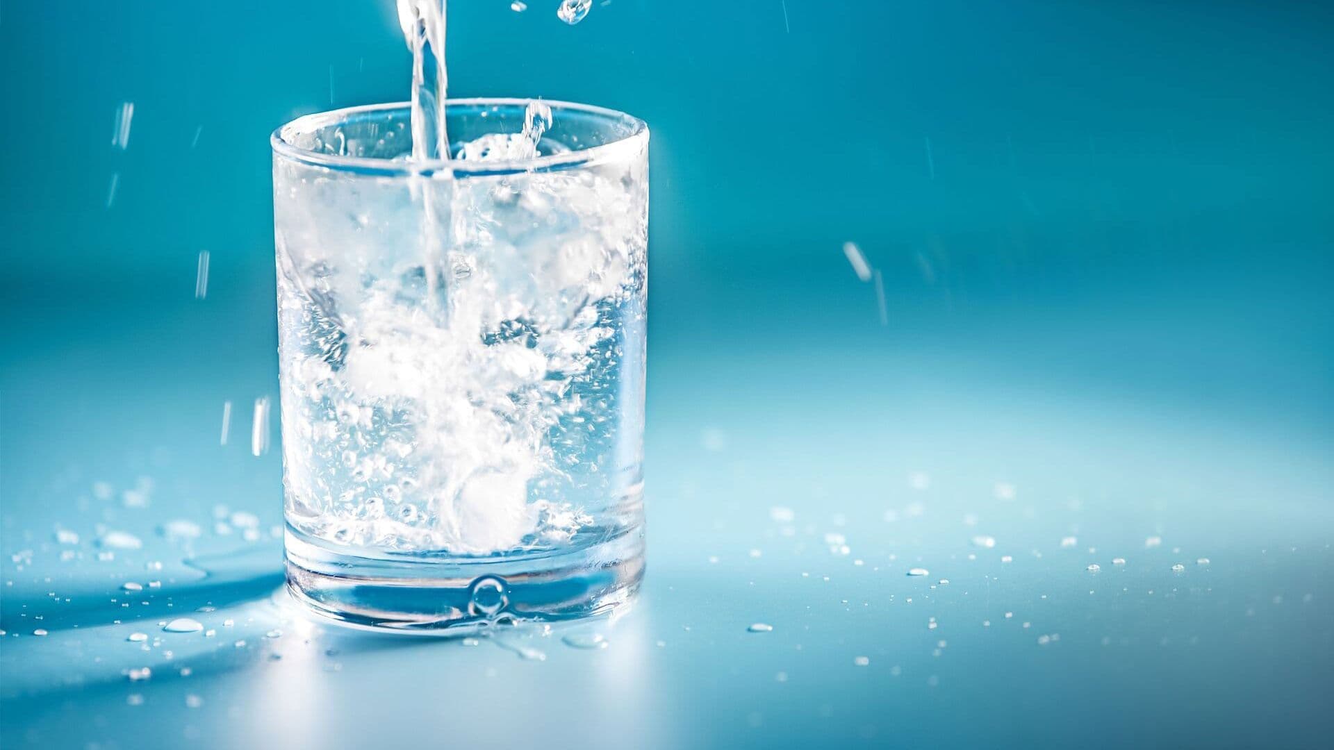 A clear glass is filled with splashing water against a bright blue background. Droplets surround the glass, creating a sense of freshness and purity.