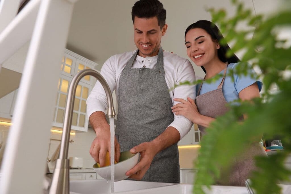 A man and a woman, both smiling, wash dishes together in a bright kitchen. He wears a gray apron, and she gently embraces him from the side.