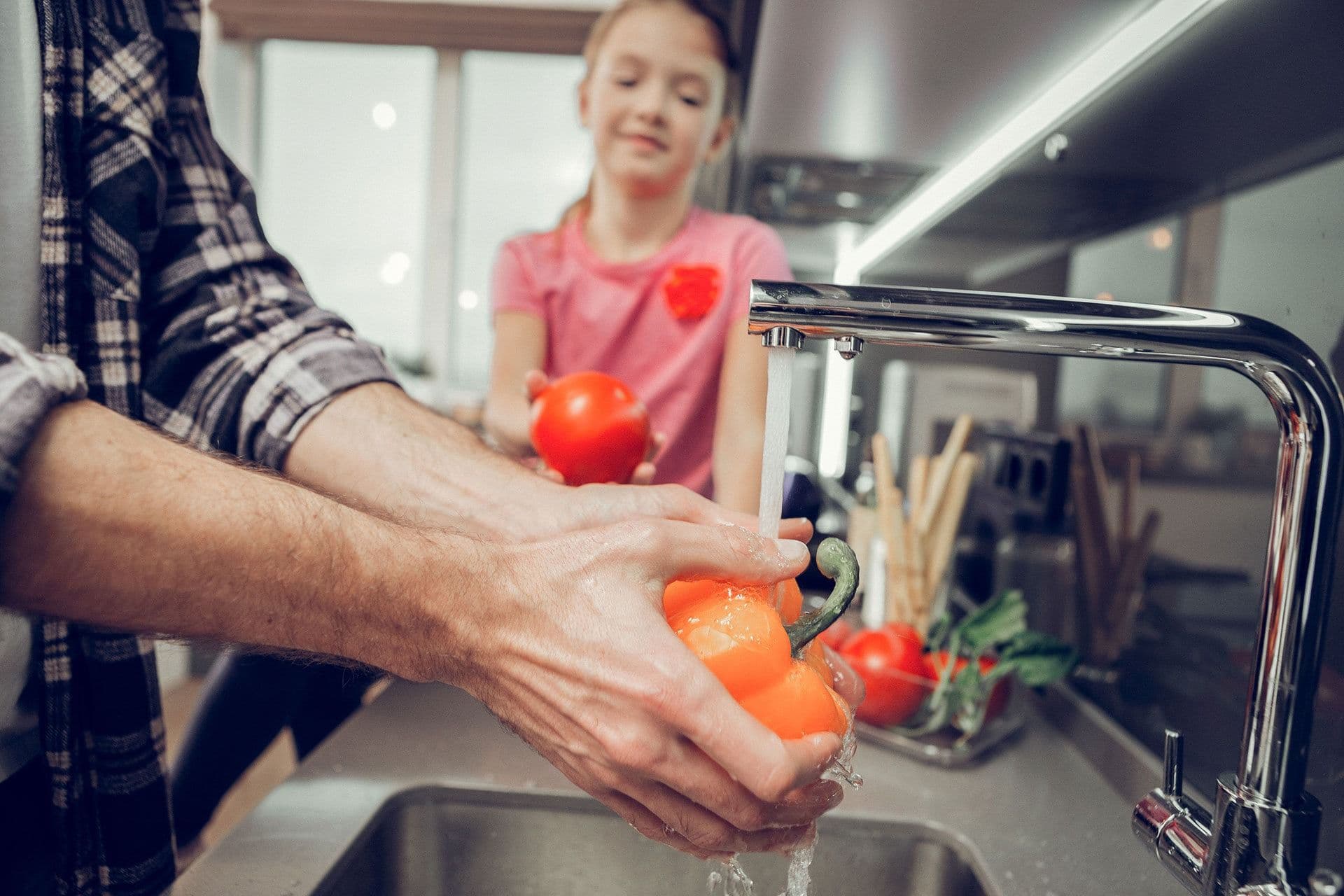 A person washing tomatoes under a kitchen faucet, with a child in a pink shirt holding a tomato and watching. The scene is bright and casual.