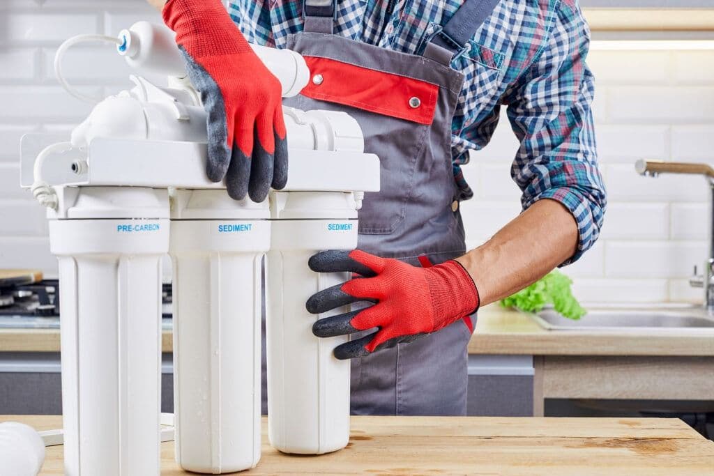 A person wearing red gloves and a plaid shirt installs a water filtration system in a kitchen. The background features white subway tiles and a sink.