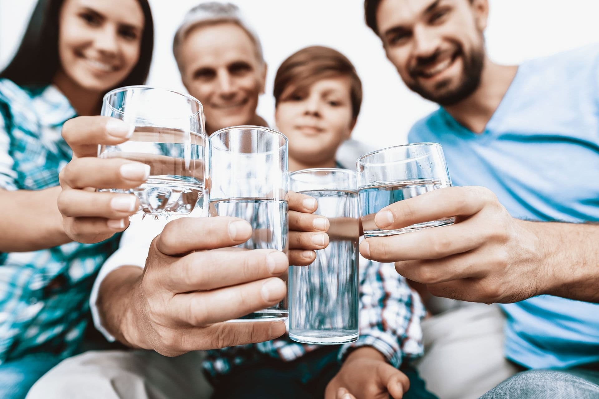 A group of four smiling people holds up glasses of water in a cheerful toast, suggesting a moment of celebration or togetherness indoors.