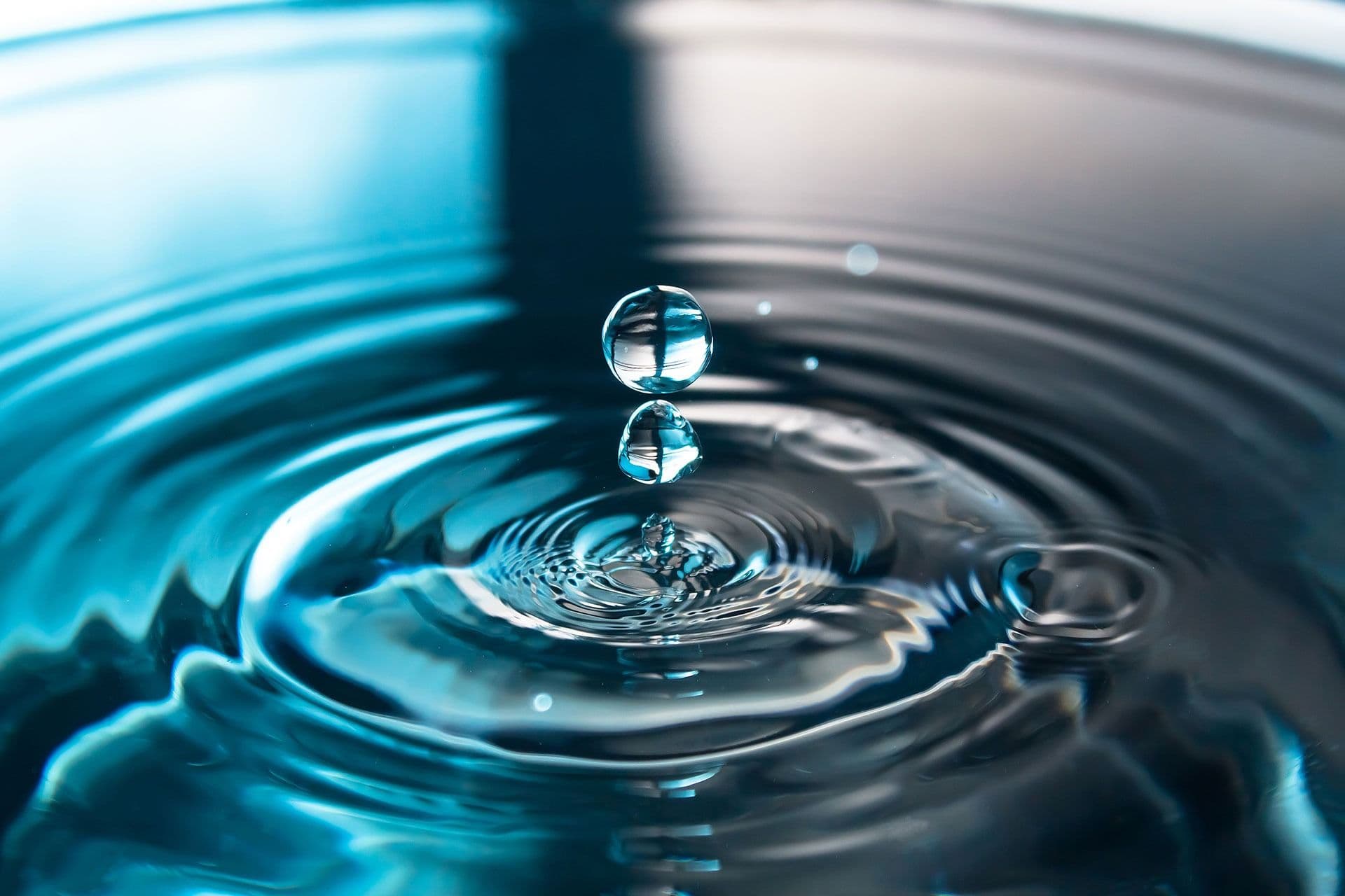 A clear water droplet captured in mid-air above a calm blue water surface, with gentle ripples forming.