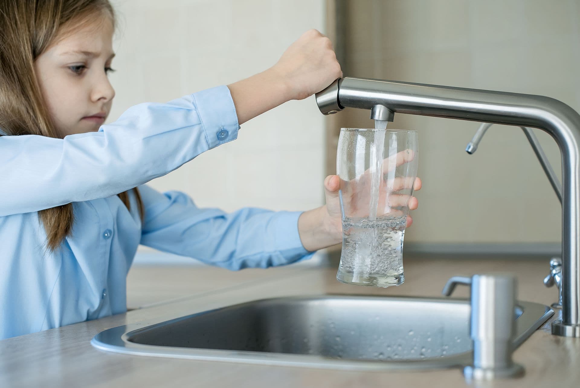A girl in a light blue shirt fills a glass with water from a kitchen faucet, focusing intently. Modern, clean kitchen setting, conveying a sense of responsibility.