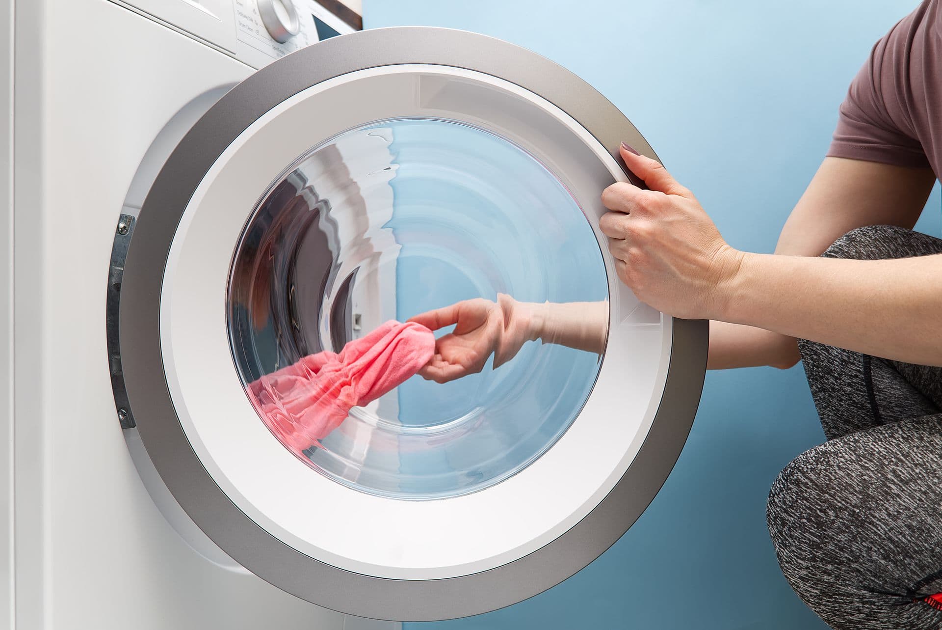 A person in gray leggings opens a front-loading washing machine, reaching for a pink garment inside. The machine is set against a light blue wall.