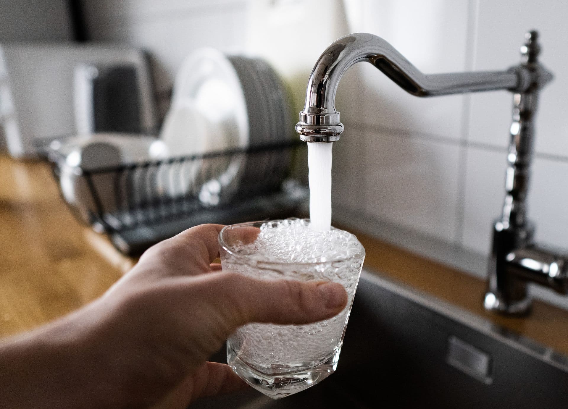 A hand holds a glass being filled with water from a chrome kitchen faucet. In the background, clean dishes are stacked on a drying rack.